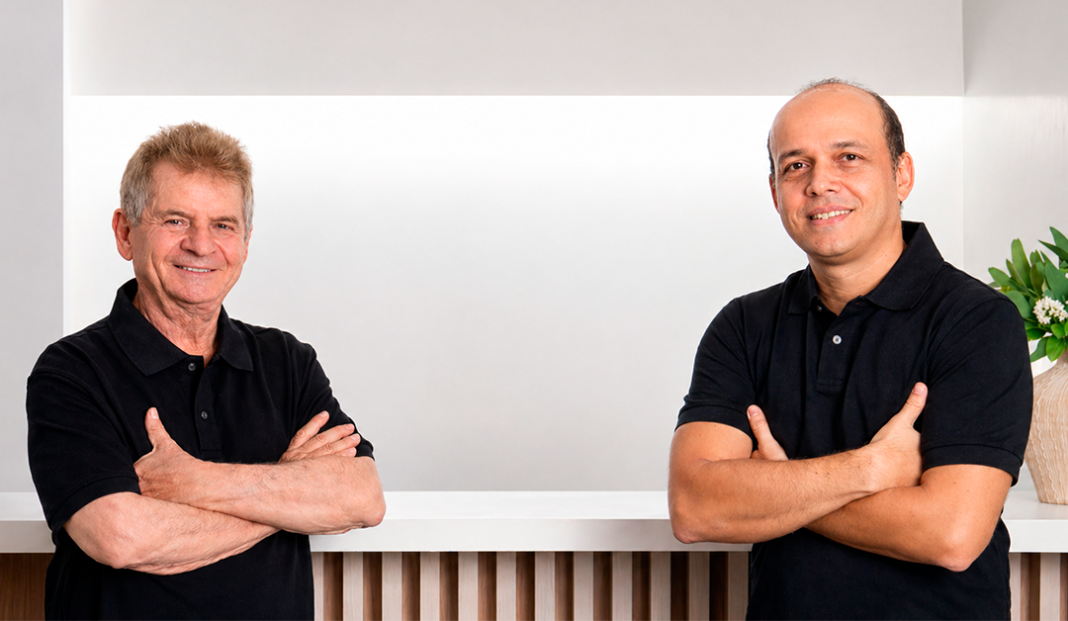 Two men with arms crossed stand behind a white counter in a bright, minimalist space, wearing black polo shirts.