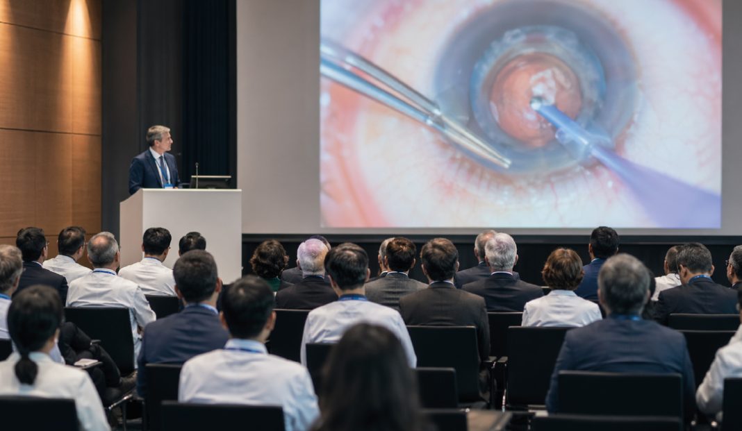 Speaker at a podium presenting to a seated audience in a conference hall, with a large screen showing an eye surgery close-up behind him.