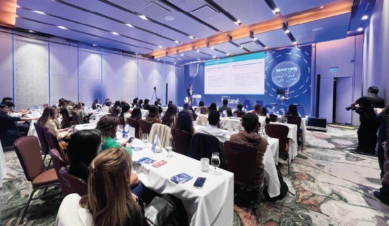 Conference room with attendees at white-clothed tables facing a stage and large slide screen at a Master Day event