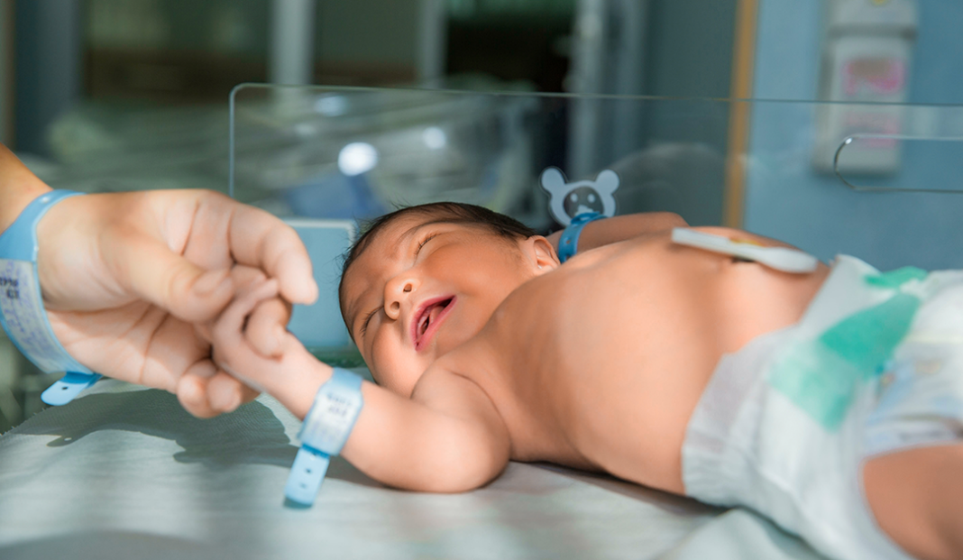 Newborn baby on a hospital bed, eyes closed, holding an adult's finger with medical wristbands visible on both.