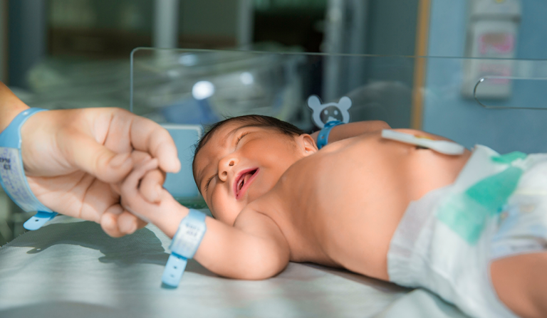 Newborn baby on a hospital bed, eyes closed, holding an adult's finger with medical wristbands visible on both.