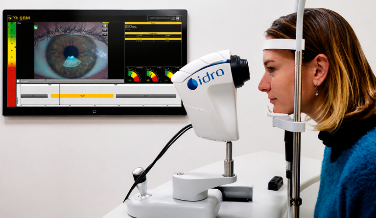 Woman undergoes an eye exam with a diagnostic instrument and a monitor showing an enlarged eye image and test data, on the wall.
