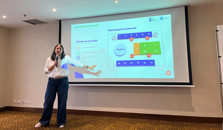 Female presenter standing at the front of a conference room, holding a microphone and pointing to a projected slide with a seating plan and investment details.