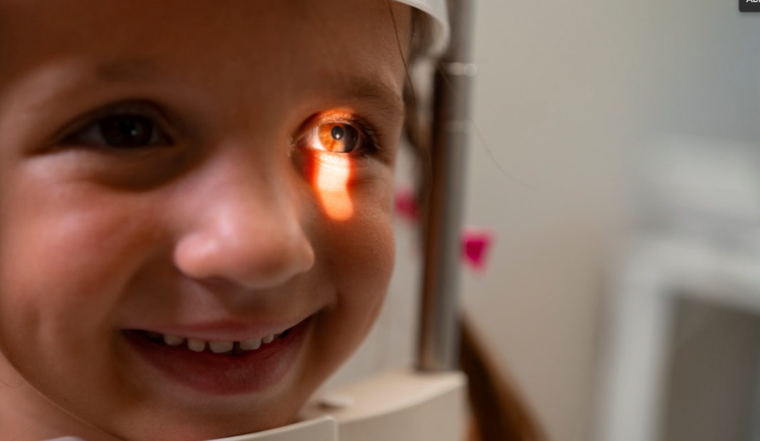 Close-up of a smiling child at an eye exam, with a light reflecting in one eye and eye‑scan equipment in the background.